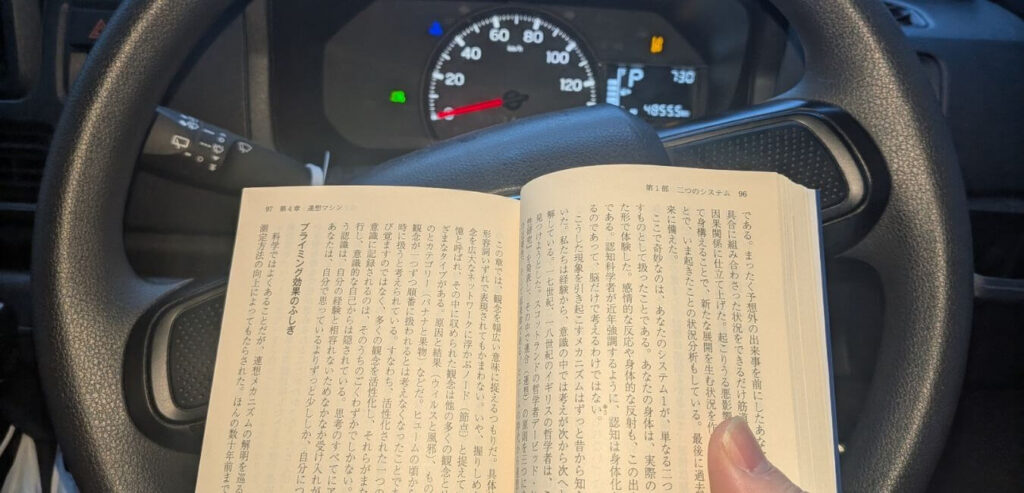 朝5時40分、職場の駐車場での読書風景：
車の中で『ファスト&スロー』を読む手元。
ダッシュボードの時計が7時台を示している。
朝日が窓から差し込む。毎日20分の積み重ねが人生を変えた。